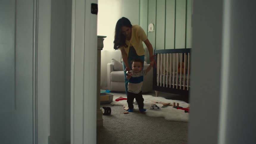 Woman guides baby from behind as they both walk towards the door