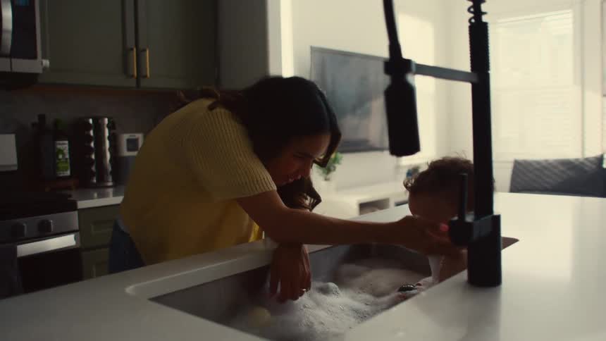 A mother is bathing her infant son in the kitchen sink