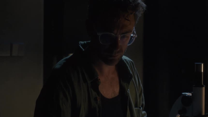 A man with glasses sits at a table looking into a microscope and then stops and looks thoughtful