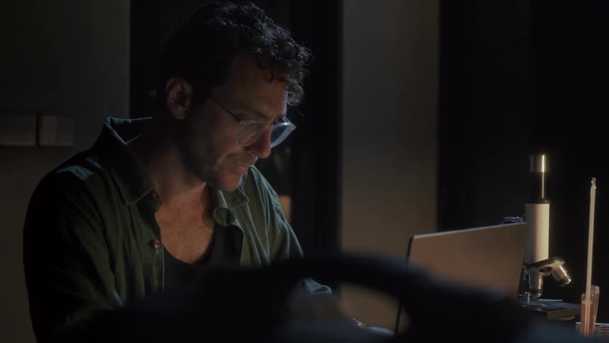 The face of a man wearing glasses who is working on a laptop while sitting in a partially illuminated room