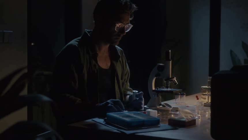 Man wearing gloves handling laboratory equipment at a table in low lighting