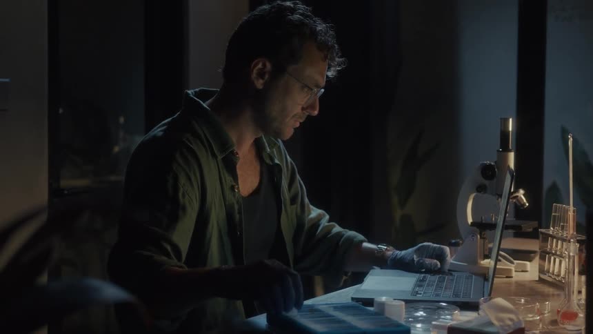 A man is using his laptop and looking at microscope slides at night in the lab