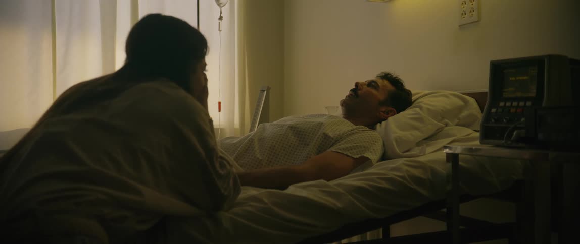 A woman sits beside a man lying in a hospital bed holding his hand in a quiet emotionally supportive moment