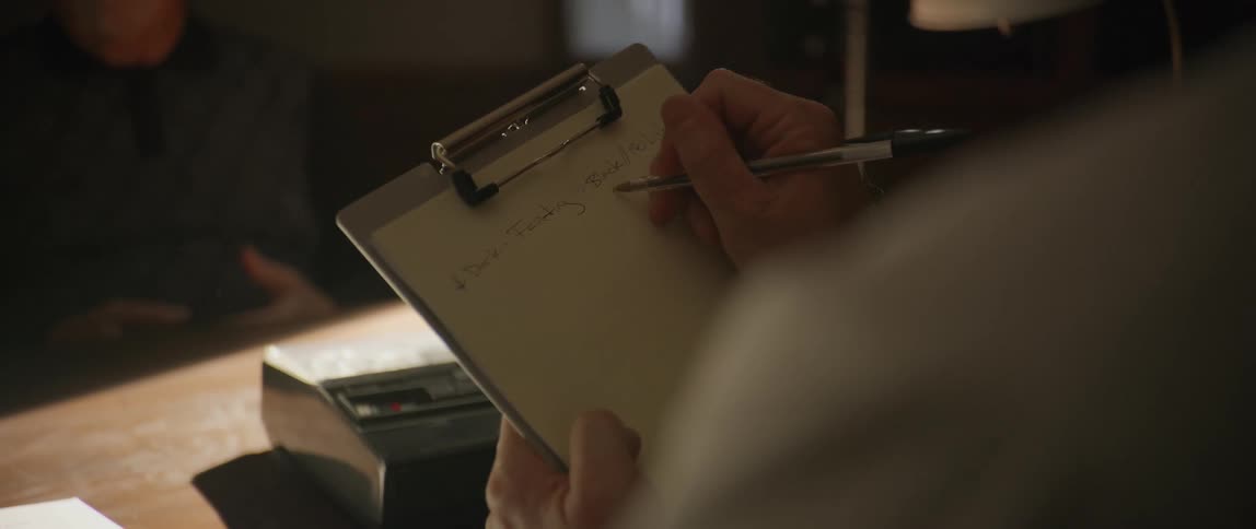 A person writes on a clipboard while another person sits on the other side of the desk