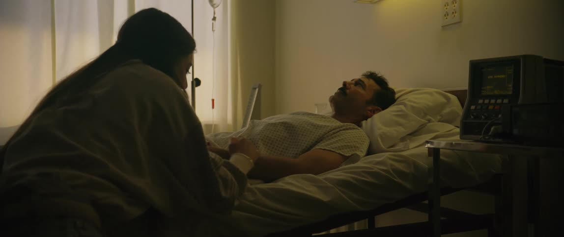 A sick man lying in a hospital bed with a woman sitting beside the bed