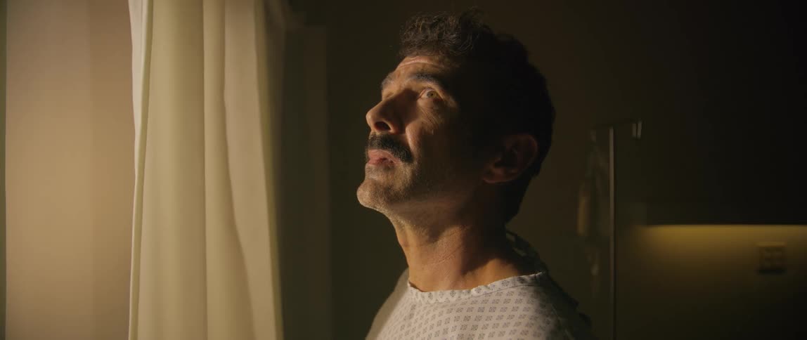 A man appearing weary and contemplative gazes out a window in a dimly lit hospital room