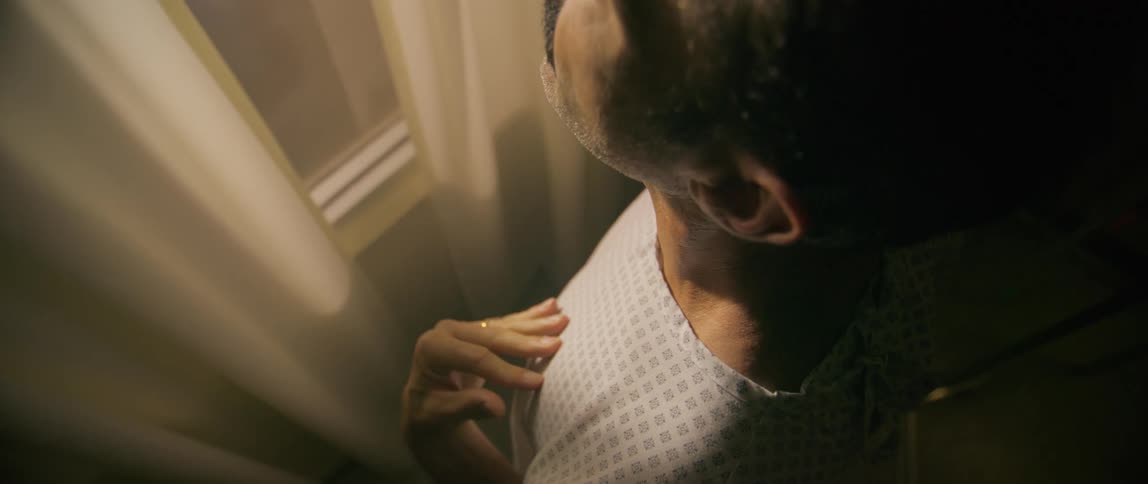 Male patient stands near the window during the daytime and looks around after touching his neck