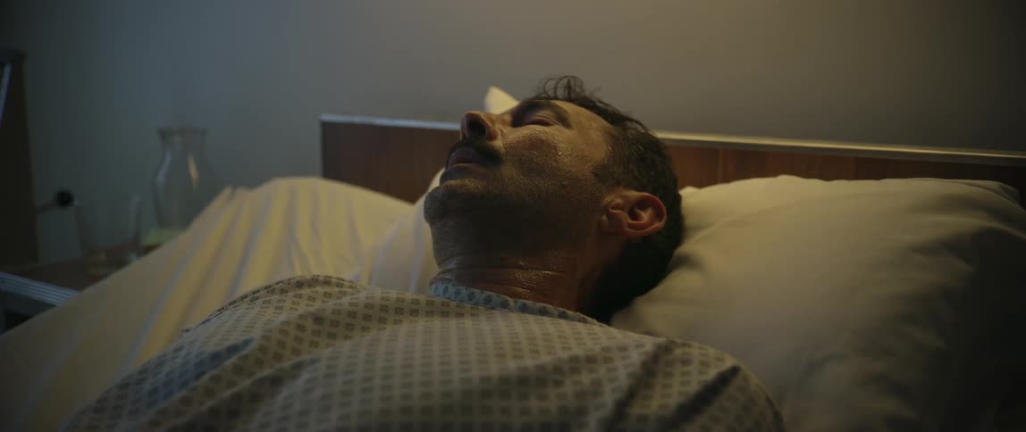 A middleaged man lies unconscious or asleep in a hospital bed under dim lighting suggesting a medical or emotional situation