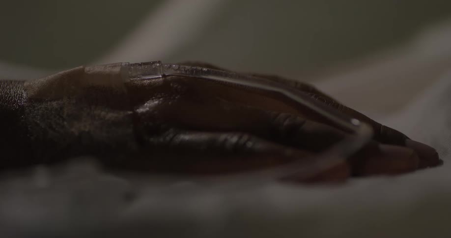 A closeup shot of a hand with an iv line inserted lying still on a hospital bed under dim lighting