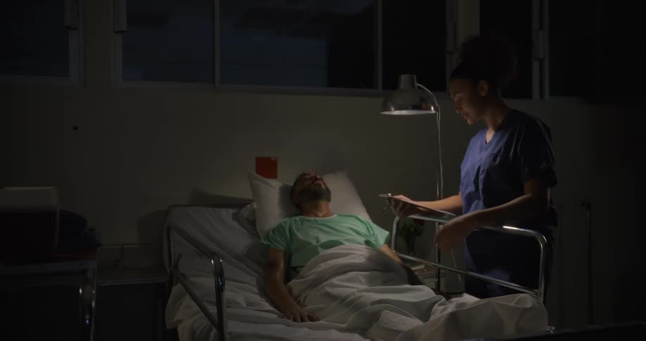A sick man sweating in a hospital bed with a nurse approaching to check on him