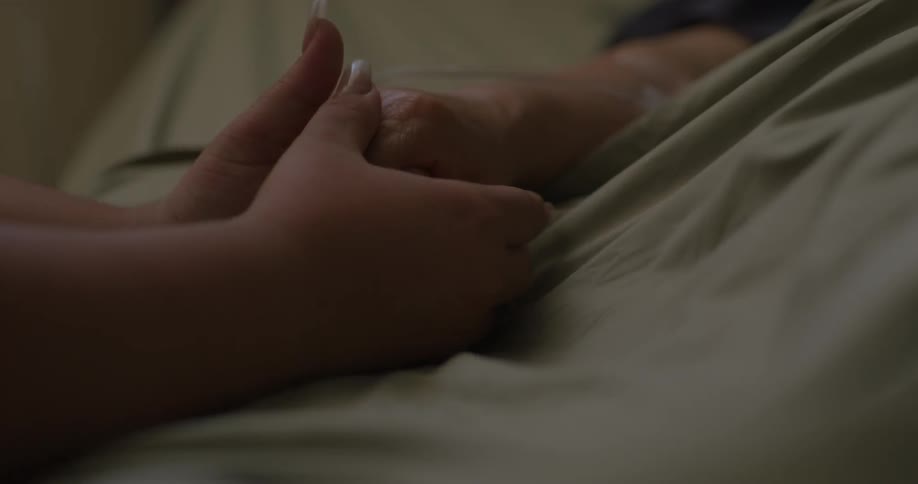 A woman holds and massages the hand of an elderly person lying on a bed