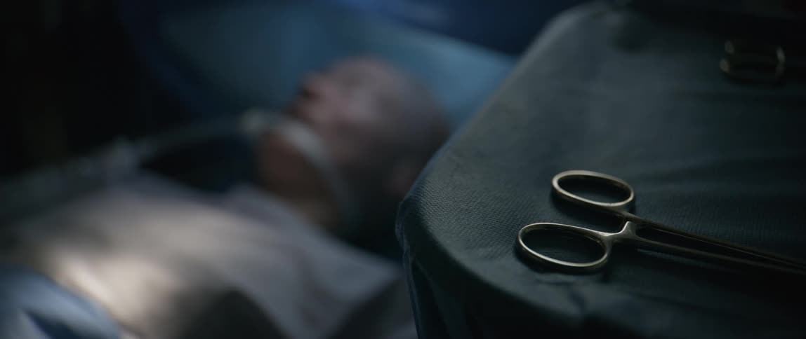 A pair of surgical scissors is focused in front of an elderly man on a respirator in a hospital bed breathing shallowly as a hand appears above him and indicates something out of frame with its index finger