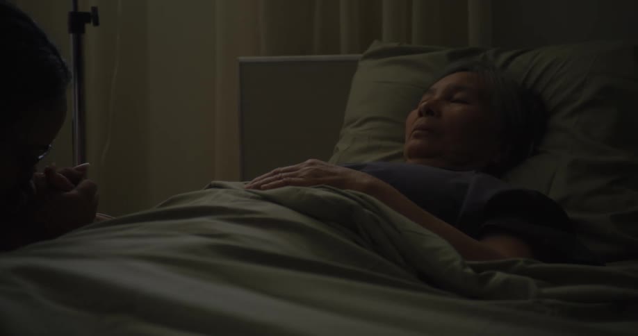 A person provides comfort to a woman lying motionless in a hospital bed within a dimly lit room