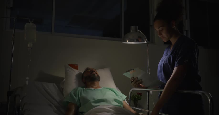 A nurse checks on a male patient in a dimly lit hospital room at night