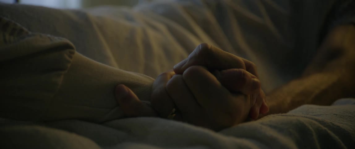 Two people hold hands in a dimly lit hospital bed conveying a sense of fear vulnerability and emotional connection in a medical setting