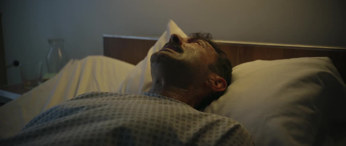A middleaged man lies in a hospital bed under dim lighting appearing distressed or unconscious suggesting a serious medical condition or emotional moment