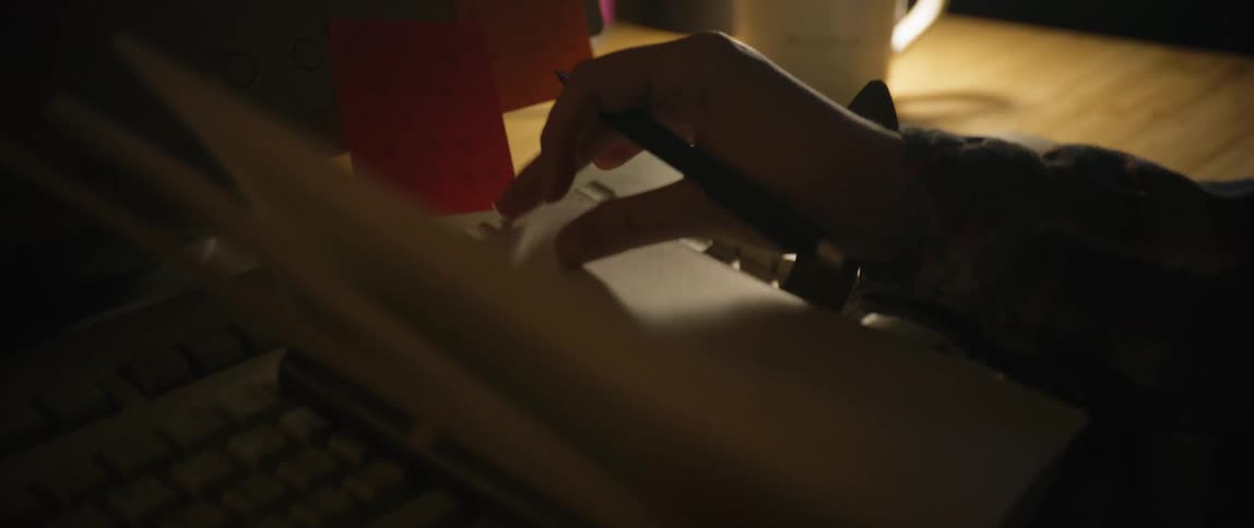 Woman sitting at her computer grabs her journal and a pen and begins to write the room is dimly lit while desk lamp is on