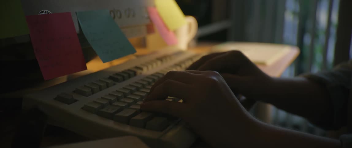 A person is typing on an older computer that has notes stuck to it
