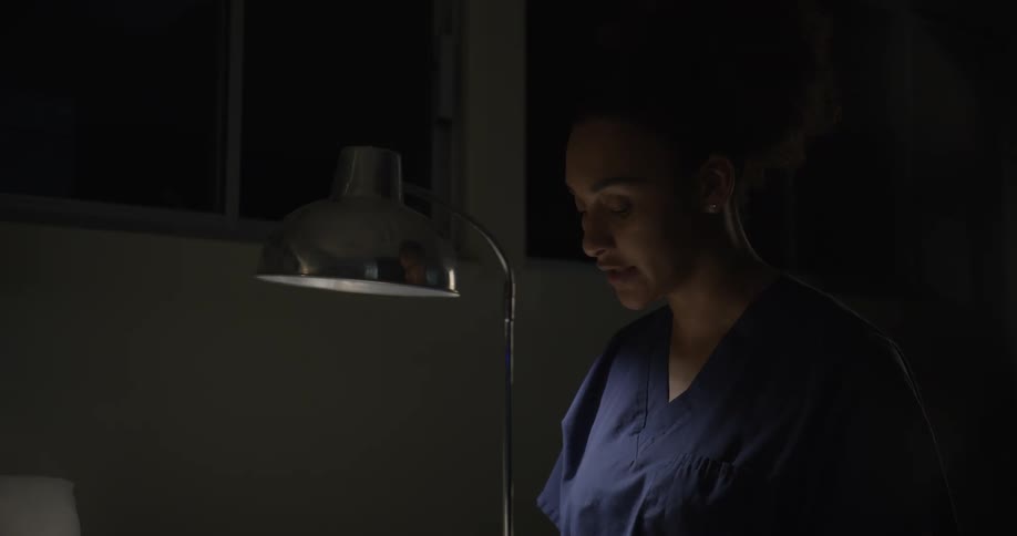 A woman in medical scrubs stands under a desk lamp seemingly engaged in a conversation in a dimly lit room