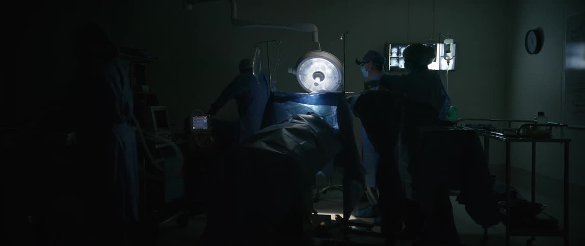 Medical team performs surgery in a dark room with light focused on the patient lying on the operating table