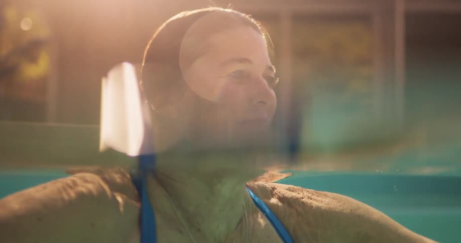 A woman joyfully swims in a sunlit pool with her smiling face emerging from the water as sunlight reflects warmly around her
