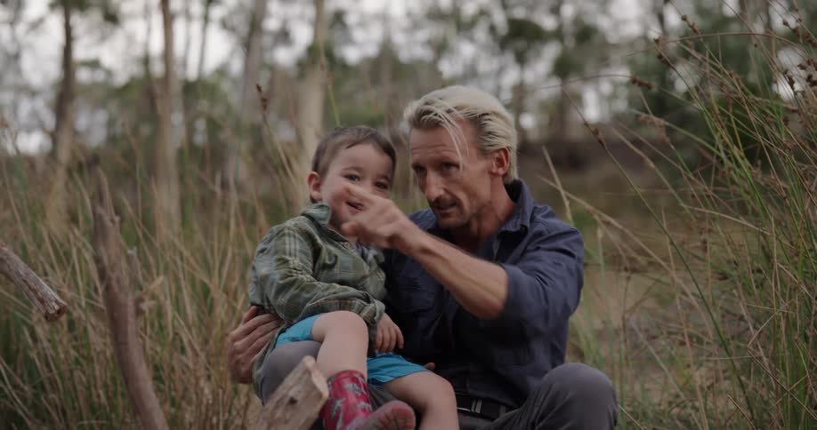 A father with blonde hair and a blue shirt bounces a young boy son wearing green on his knee while laughing and pointing near the camera