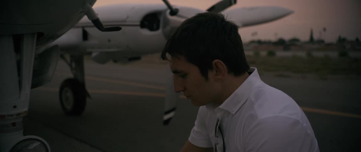 A young man possibly a pilot or technician is seen beside a small aircraft on a runway during dusk appearing focused or deep in thought