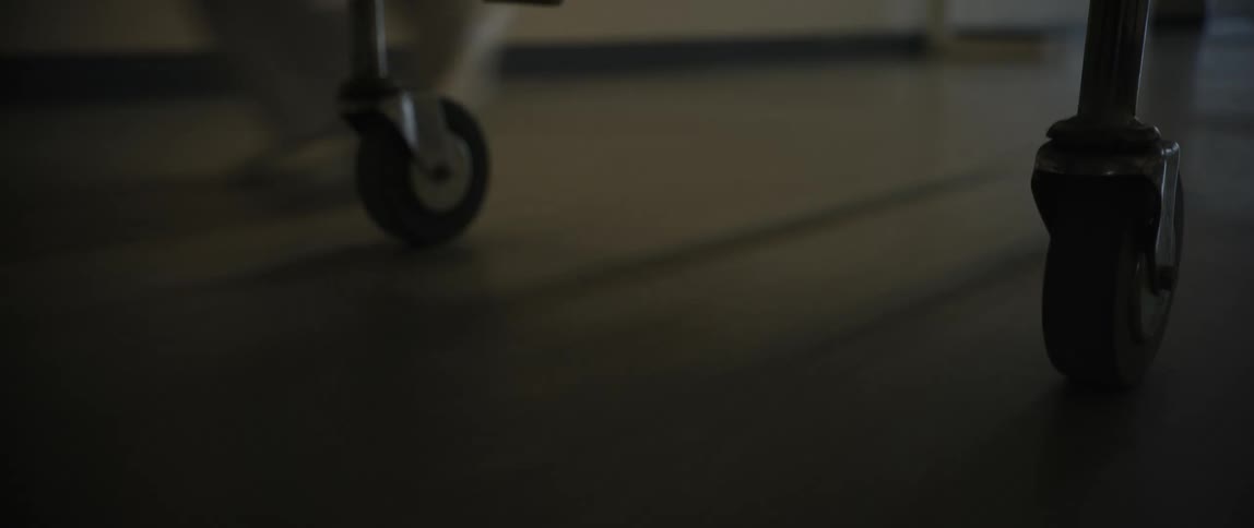A closeup shot of a rolling cart or medical trolley being pushed along a dimly lit indoor floor with no visible faces or identifiable people