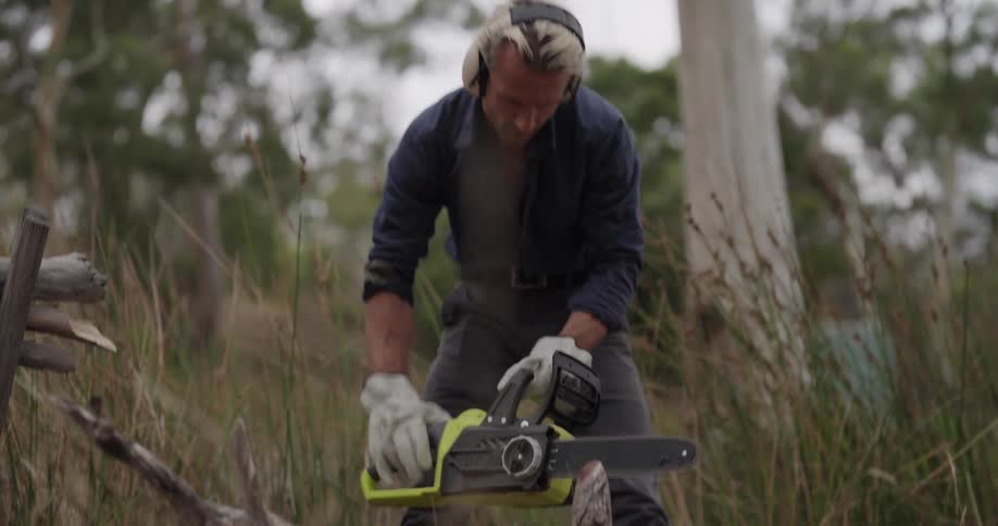 A man in protective gloves is using a chainsaw to cut large tree branches in a grassy outdoor setting