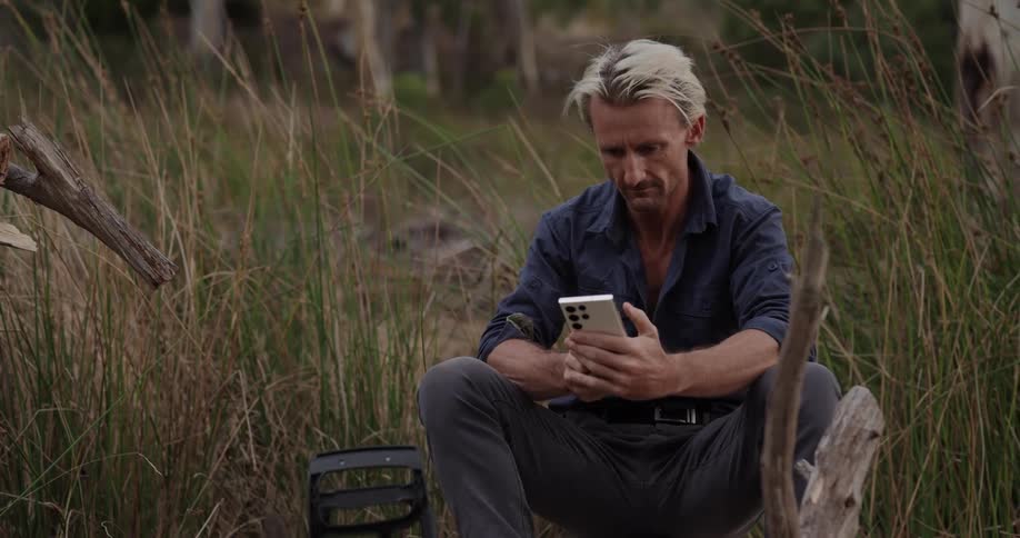 Man sitting in a field with tall wheat grass texting on cell phone