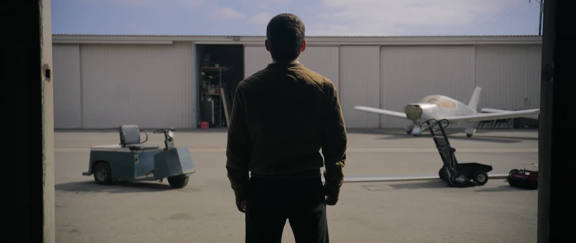A warehouse door opens and a man wearing glasses looks out with a view of an airplane in the background