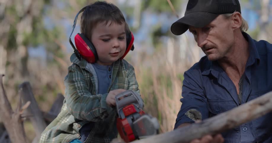 Boy with a toy chainsaw cutting a piece of wood accompanied by his father