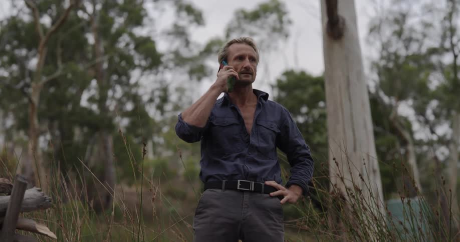 Man answering a phone call while standing in an outdoor setting surrounded by trees