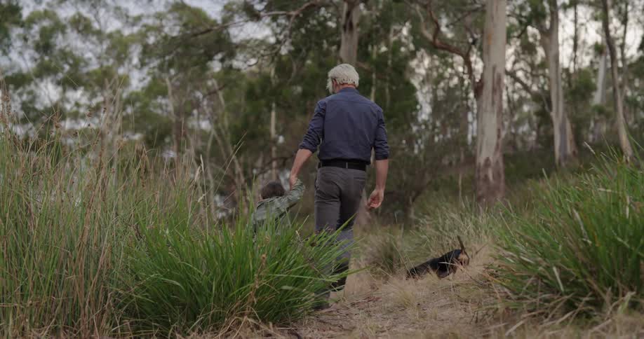 A father and his young son walking hand in hand through a wooded area with their dog beside them