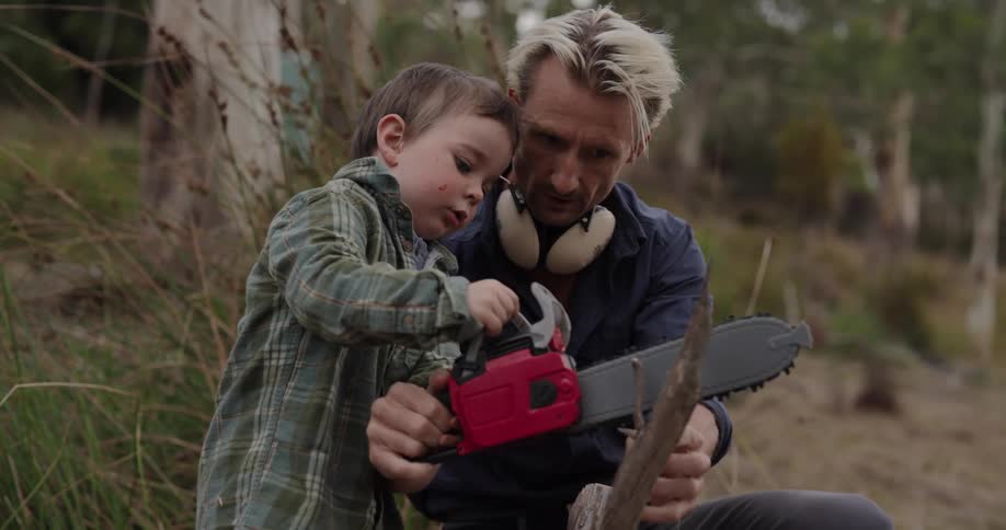 A grizzled man demonstrates the use of a chainsaw to his young son amidst a rugged wooded landscape