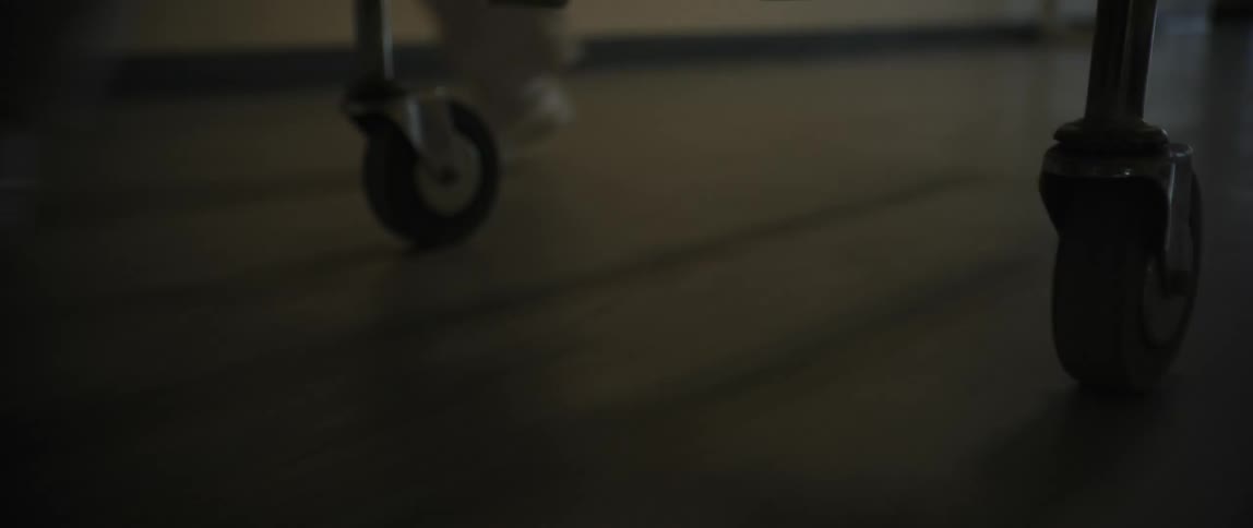 A hospital gurney is being wheeled down a dim hallway with a person walking alongside it seen from a low closeup angle of the wheels