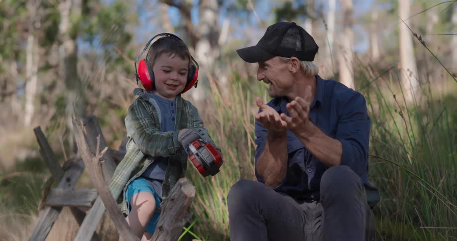 A father and his son outdoors near trees playing with a toy chainsaw and a piece of wood