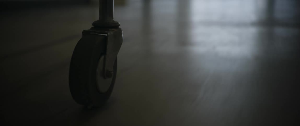 A wheel of a gurney is moving across a polished floor in a dark hallway