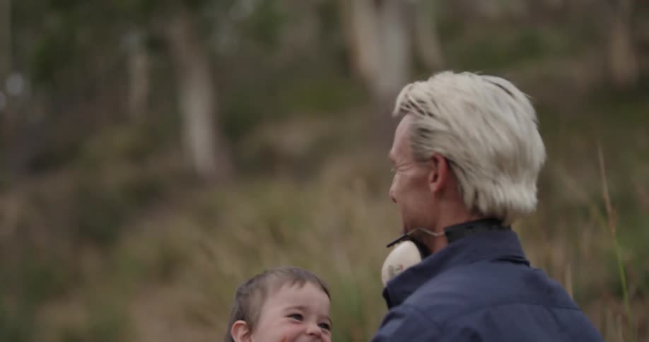 A father with very light hair is wearing head phones around his neck and and has a blue jacket tosses his son who is wearing a green plaid jacket in the air and catches him the young boy is having fun and laughing as he is being tossed in the air