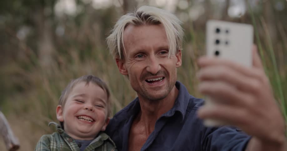 A father and son wave at the cell phone while taking a selfie