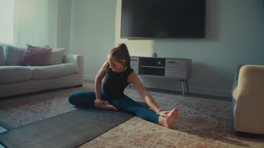 Young woman stretches while sitting on a yoga mat in her living room