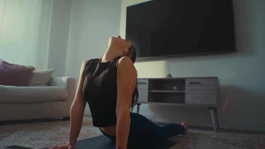 A young woman with long hair in a ponytail and wearing a tight fitting black halter top and pants is doing a yoga pose on a yoga mat in a clean sparse living room with a big screen tv and white sofa with white curtains closed over a large window