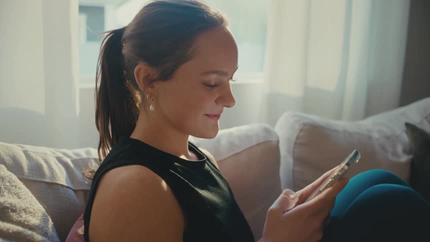 Woman smiling texting on cell phone sitting on couch at home then anxiously looking around