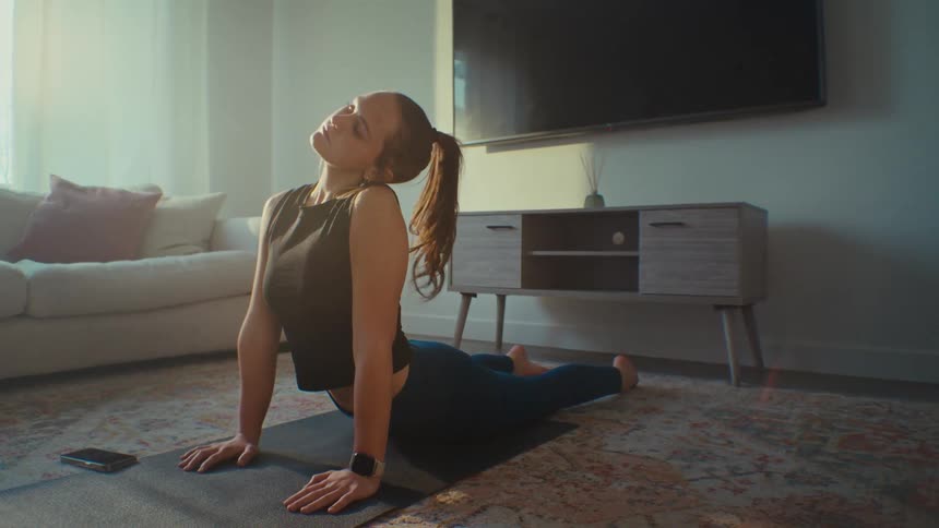 A young blonde woman is doing pilates in a living room and then starts using her cell phone