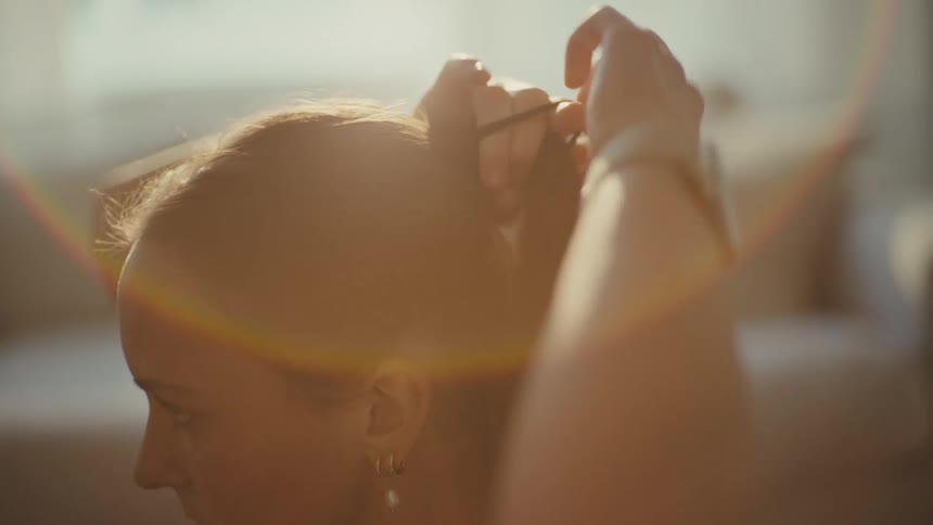 A young woman sits in a softly sunlit room tying her hair back in a calm contemplative moment