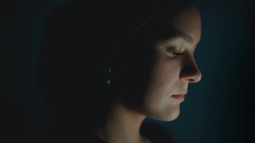 A young woman is quietly illuminated in profile eyes closed and expression calm suggesting introspection or focus in a dim interior setting
