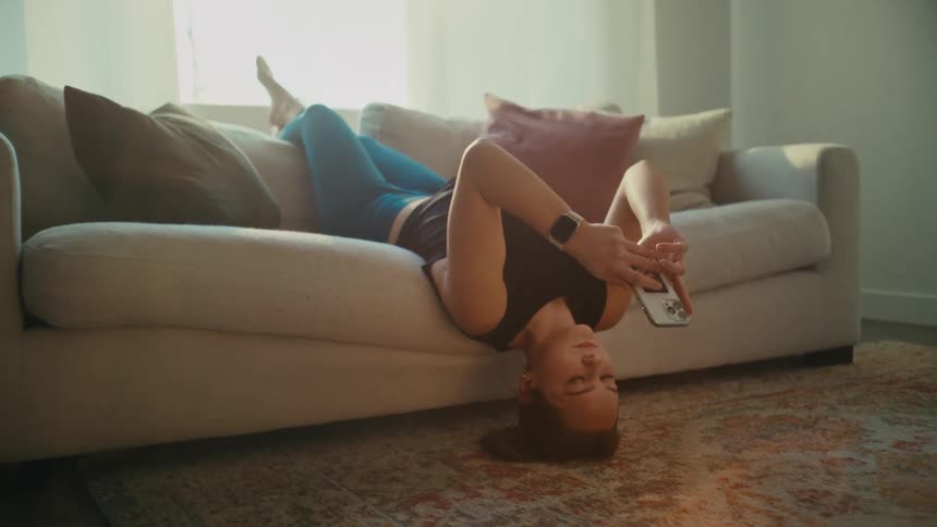 A woman lays upside down on her couch while looking at her phone