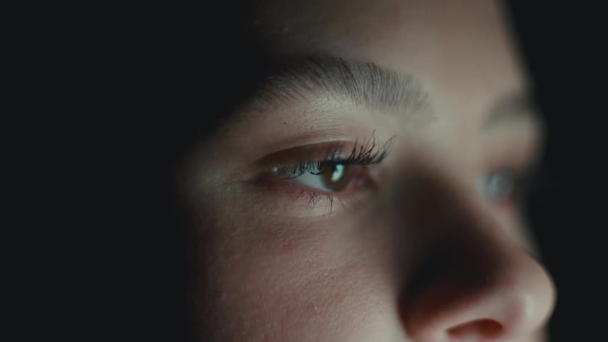 A young woman blinking while watching a computer monitor at night