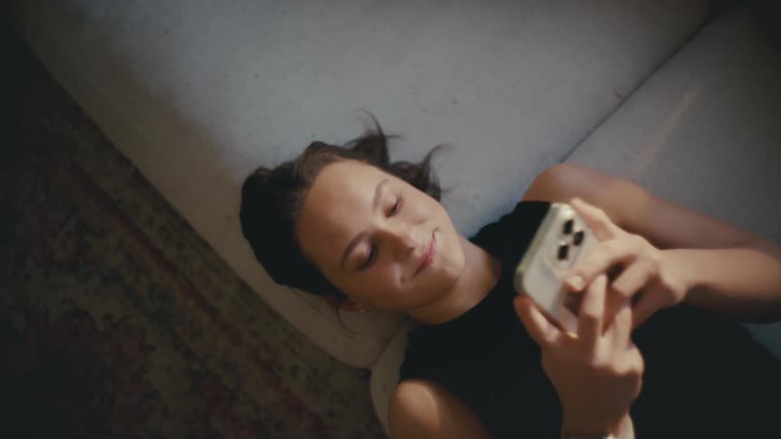 A teenage girl lays on a couch happily texting on her smart phone