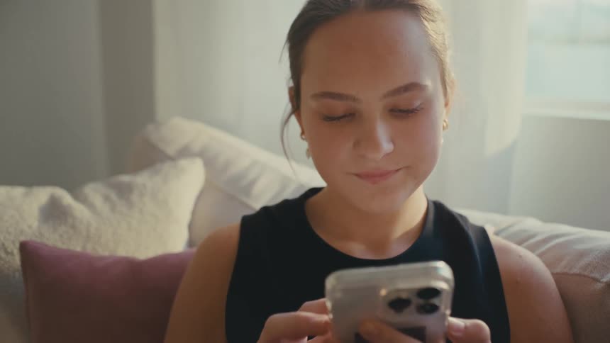 A young woman smiles as she uses her phone on a sofa